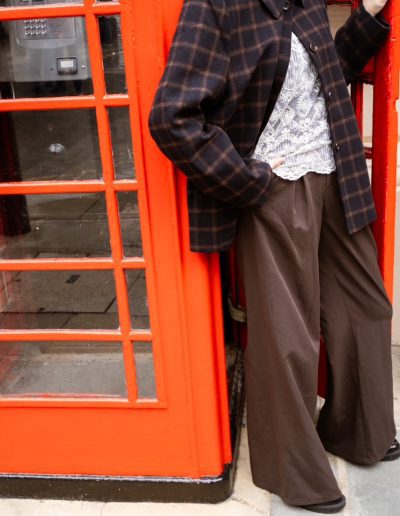 Plaid overshirt, lace top and wide-leg brown trousers, leaning in the open doorway of a red phone box