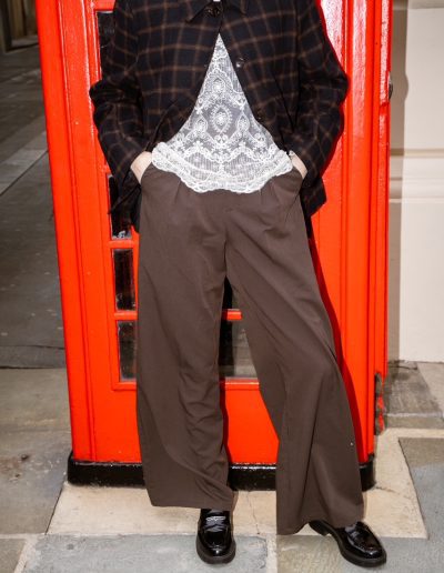 Plaid overshirt over a lace top with wide-leg brown trousers and loafers, hands in pockets, in front of a red phone box