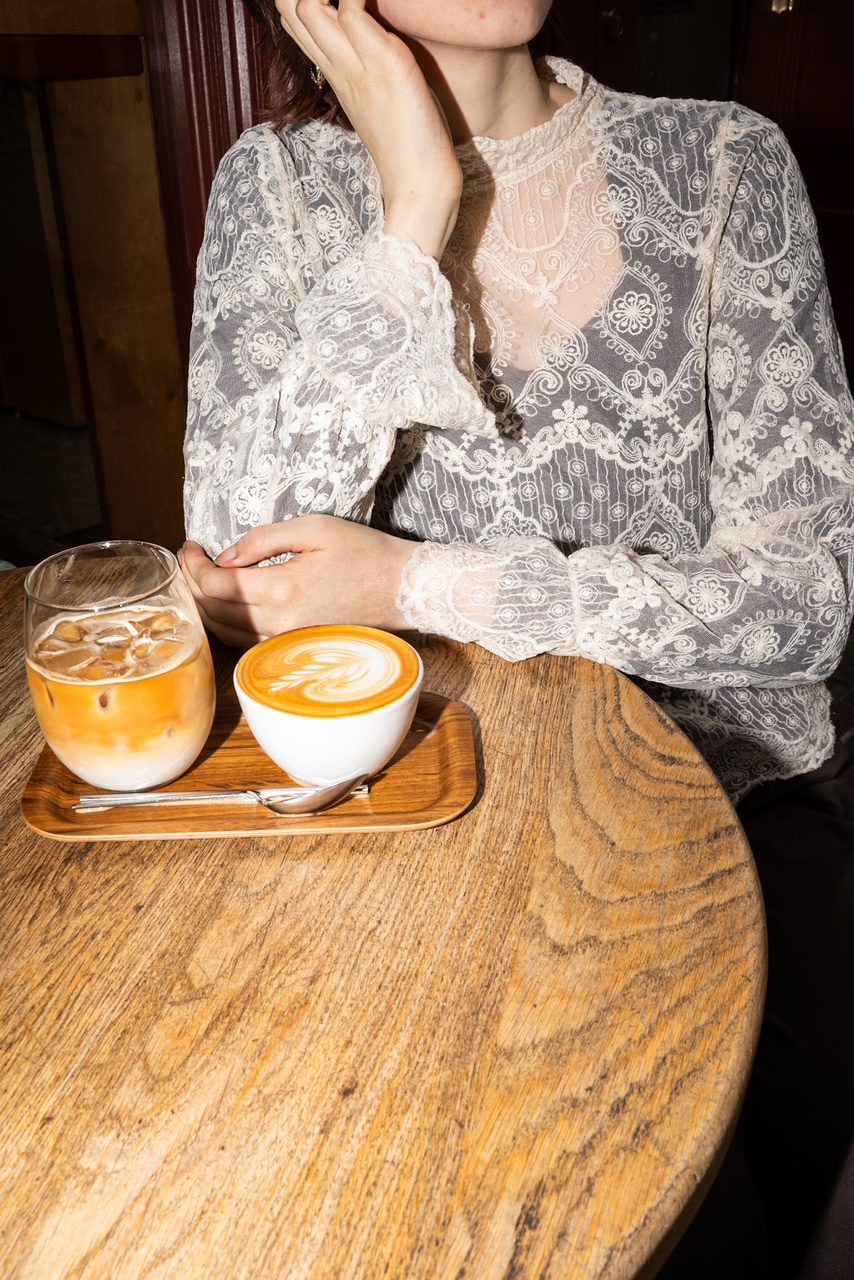 Lace blouse at a wooden cafe table with an iced latte and a cappuccino